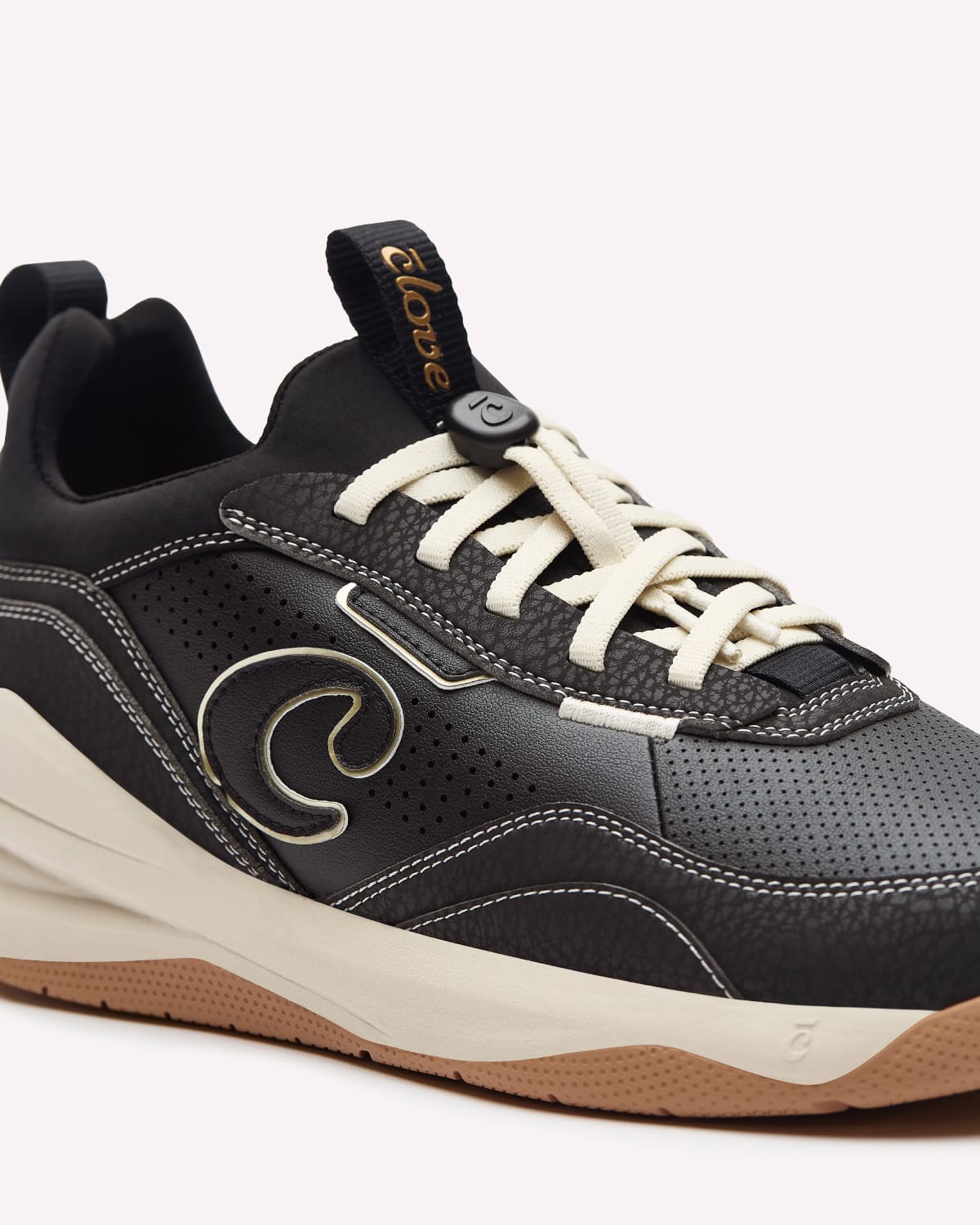 A close-up of a black and cream sneaker with a brown gum bottom, featuring a textured leather upper, cream laces, and a decorative gold-tone logo on the side.