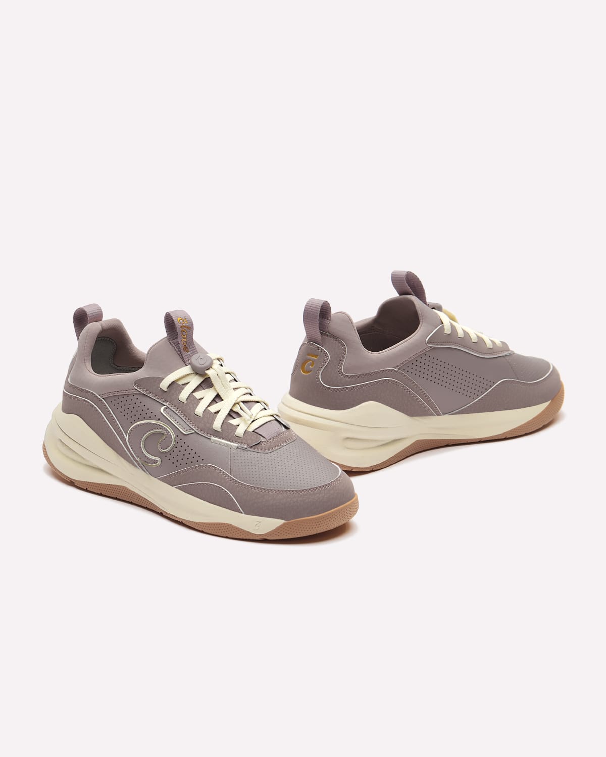 A pair of low-top sneakers in a muted taupe-gray colorway with cream laces and a cream mid-section, accented by a small gold logo on the side and heel.