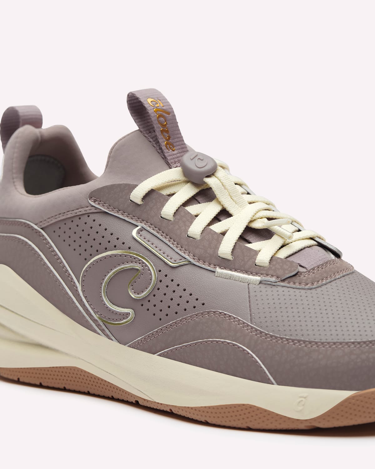 A close-up of a sneaker in taupe and light gray with cream laces and a brown bottoms, shown against a light off-white background.