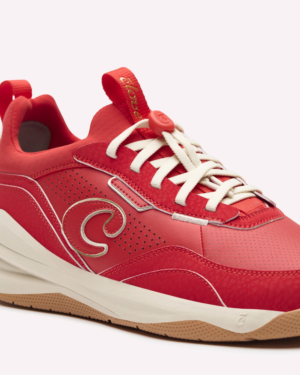A red athletic sneaker with off-white laces, a cream mid-section, and a gum bottom, featuring a large stylized 'S' logo on the side and a pull tab with gold lettering.
