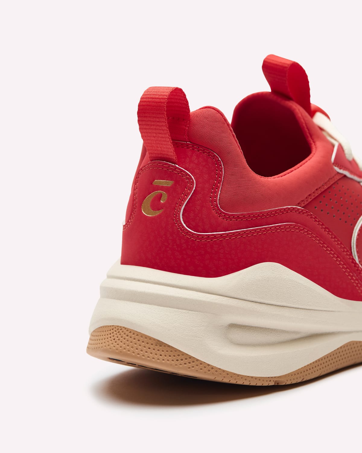 A close-up rear three-quarter view of a red low-top sneaker with a thick cream mid-section and a gum bottom, featuring a gold logo on the heel.